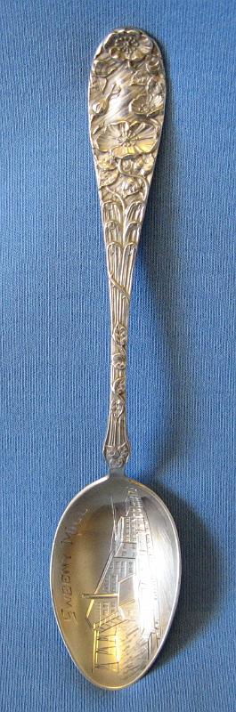 Souvenir Mining Spoon Sweeny Mill.JPG - SOUVENIR MINING SPOON SWEENY MILL WARDNER ID - Sterling silver spoon withengraved bowl showing mine buildings and marked SWEENY MILL with figural handle of flowers, 5 3/4 in. long, reverse marked Sterling with maker’s hallmark  (The Coeur d'Alene mining region in northern Idaho is one of the nation's most significant mineral producers, having yielded almost twenty percent of the silver and lead, and six percent of the zinc produced in the United States.  Due to the relatively low-grade of many of the lead-zinc-silver ores mined, early methods of concentration were introduced at the beginning of commercial mining operations in 1886. These milling operations produced large tonnages of mill tailings, the material remaining after crushing, grinding of the ore and separation of a significant portion of the economic minerals as a waste product.  The Sweeny or Last Chance mill located about two miles west of Kellogg, ID in the Coeur d’Alene District was a major contributor to the production of lead and silver as well as tailings.  In 1886 Charles Sweeny and F. R. Moore of Spokane opened the Last Chance mine, located near Wardner.  In May 1898, the Empire State-Idaho Mining and Development Company was organized to control the Last Chance and to acquire new territory to the west. In September 1903, the Federal Mining and Smelting Company was organized and purchased the Empire State holdings.  The Last Chance mine was a steady producer of lead-silver ore since 1890.  With the completion of the Oregon Railway and Navigation Company's Wallace branch in 1890, Charles Sweeny built a mill with a capacity of 500 tons daily to process ore from the Last Chance mine.  At the time, nearly 300 men were employed at the mine and mill.  The Last Chance mine was worked through a tunnel nearly two miles long that perforated the ridges west of Wardner at an elevation of 3,050 feet. One portal was called the Sweeny tunnel and the other the Arizona. Concentrating ore was taken from the Arizona tunnel over the Oregon Railway and Navigation track to the Sweeny mill at the mouth of Government gulch. Lingering litigation between the Last Chance Mining Company and its successors on the one hand and the Bunker Hill and Sullivan Company on the other over ownership of the deposits was ultimately decided in favor of the Bunker Hill and Sullivan Company.  The Last Chance mine and the Sweeny mill became the property of Bunker Hill & Sullivan Mining Co. in 1918.  Mitigation of heavy metals-bearing, tailings-contaminated river sediments continues as an issue in the region today since these sediments first reached farms and communities down river from the mines around 1900.)